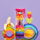 Lisa Frank Rainbow Fade BlendJet 2, portable blender, at the bottom is the Rainbow Fade colors pink, orange, yellow, green, and purple; with the blend button, with a pink cap and a little plastic pink strap on the left side of the cap, inside the blender there different kinds of fruit, at the left, there is a rainbow with a banana on the top, and on the right there a mango, a rainbow, and tree fruit hearts. 